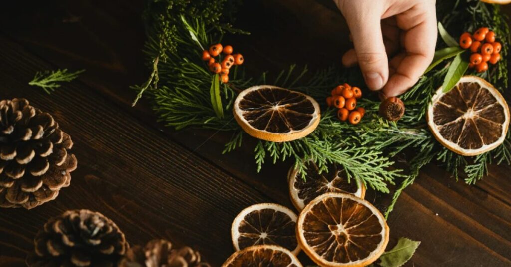 Hand arranging dried orange slices, red berries, and greenery on a wooden surface, surrounded by pinecones, creating a festive holiday wreath.