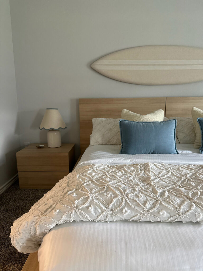 Coastal-inspired bedroom features a wooden bed, textured white bedding, blue accent pillows, and a surfboard decor piece on the wall.