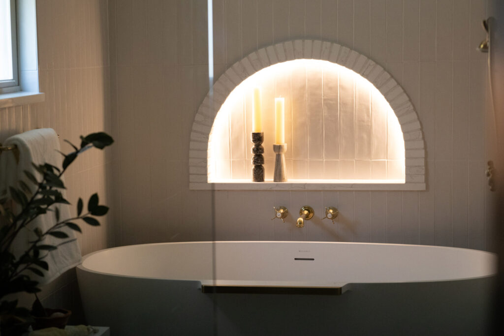 Elegant bathroom featuring a freestanding white tub, golden fixtures, and an arched alcove with candles and decorative objects.
