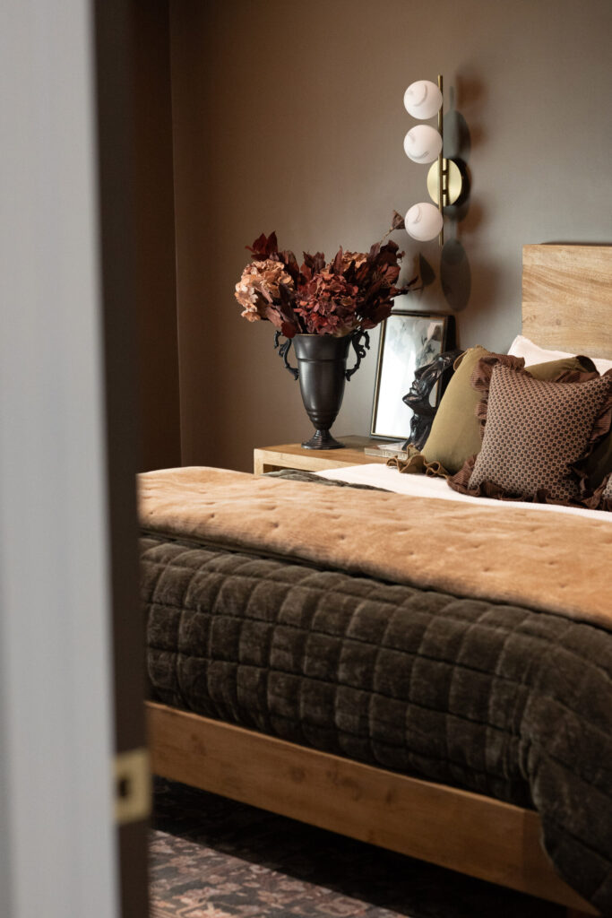 Warm-toned bedroom featuring a plush green bedspread, decorative pillows, a vase of dried red foliage, and modern wall lighting.