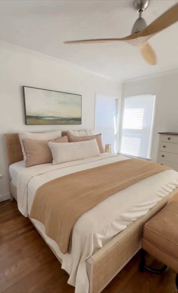 Bright, airy bedroom featuring a queen bed with neutral linens and a warm tan throw, accented by wooden floors and a ceiling fan.