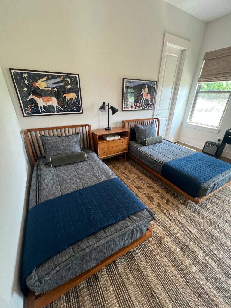 Two twin beds with gray bedding topped by navy throw blankets, a wooden nightstand with a lamp, and colorful art pieces on the walls.