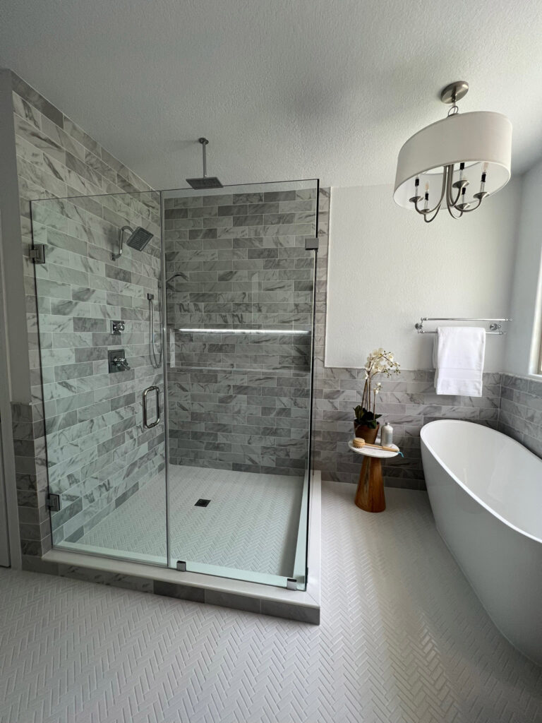 Modern bathroom featuring a glass shower with marble tiles, a freestanding bathtub, and a decorative wooden stool with plants.