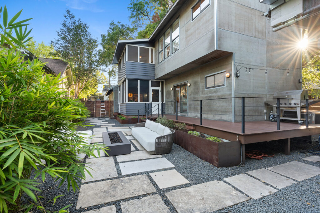 Modern backyard with a concrete and wooden deck, white sofa, stone pathways, bamboo plants, and a grill, surrounded by greenery.