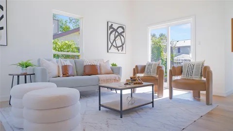 Bright living room with a light gray sofa, brown leather chairs, a round table, and large windows offering natural light.