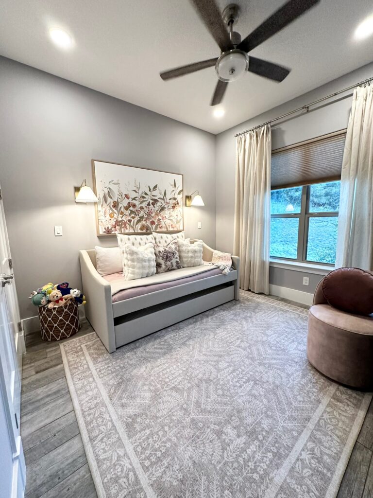 Cozy room featuring a light gray daybed with decorative pillows, a pink accent chair, and a patterned rug, illuminated by wall sconces.