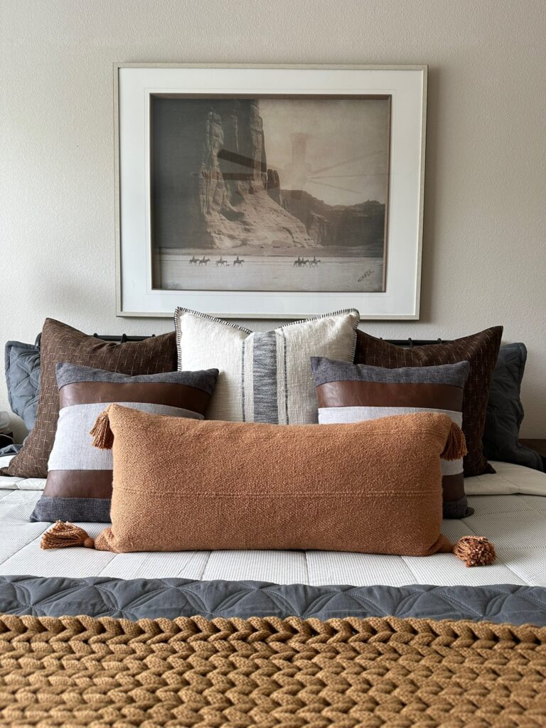 Layered pillows in earthy tones adorn a gray quilted bedspread, with a woven rug and framed landscape art above, creating a cozy atmosphere.
