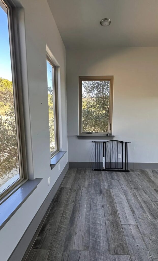 Bright study area featuring two large windows with natural light, gray wood flooring, and a black folding gate against a light-colored wall.
