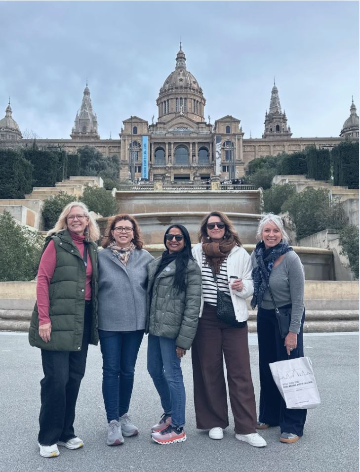 Five women stand in front of a grand historic building with domes and ornate architecture, landscaped gardens, and a cloudy sky.