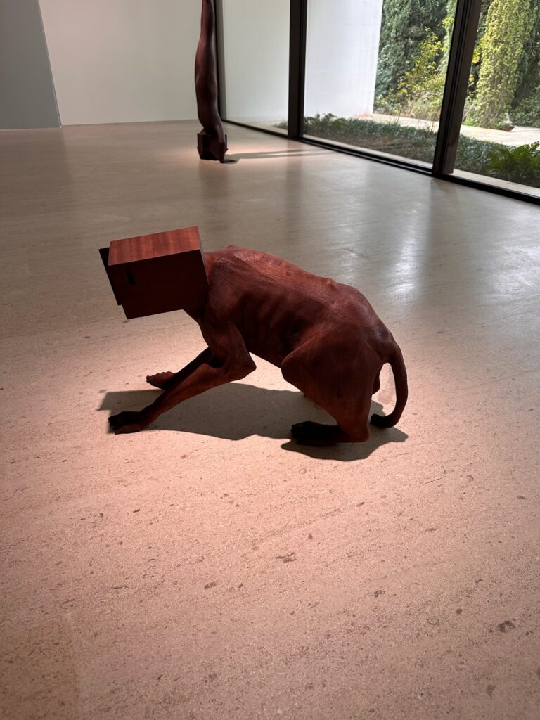 Sculpture of a dog with a wooden cubical head, positioned on a light-colored floor in a contemporary gallery setting.