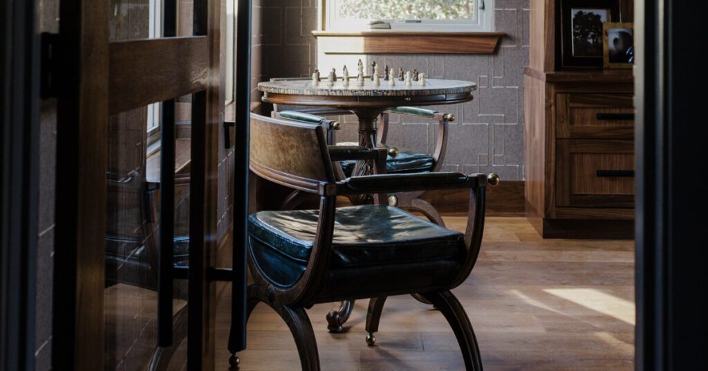A stylish wooden table with a chess set sits between two elegant chairs, bathed in warm natural light from a nearby window.