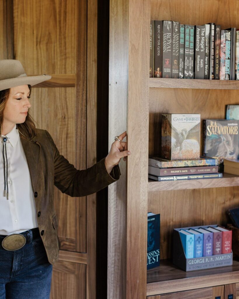 A woman in a wide-brimmed hat touches a wooden bookshelf filled with books, showcasing warm tones and a stylish interior.