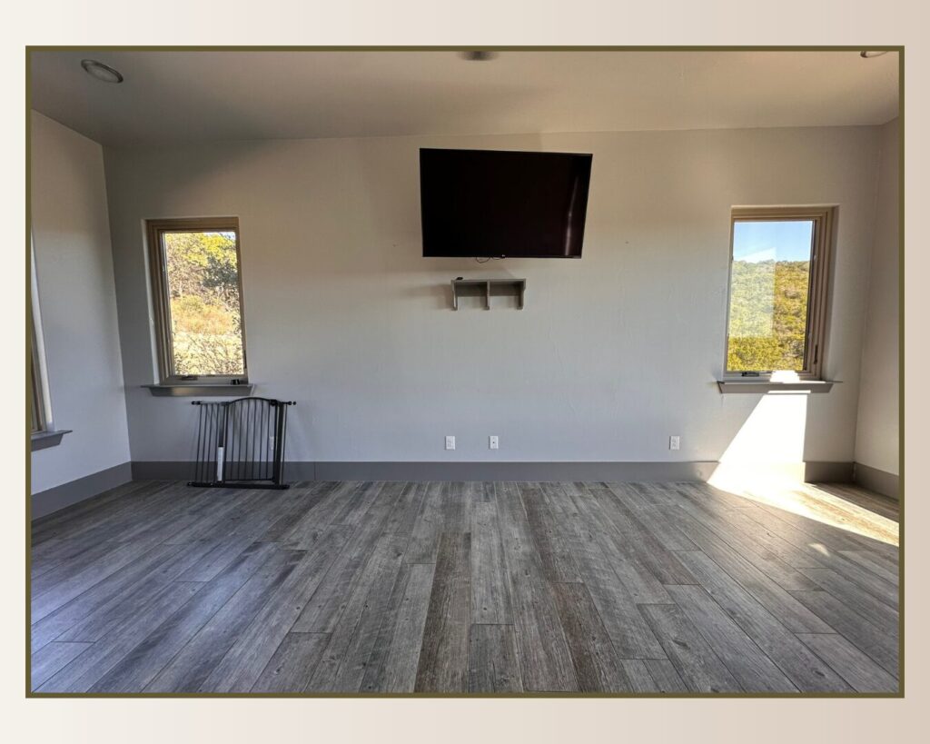 Bright, spacious interior featuring a flat-screen TV mounted on the wall, two windows, and light wood flooring, creating a modern look.