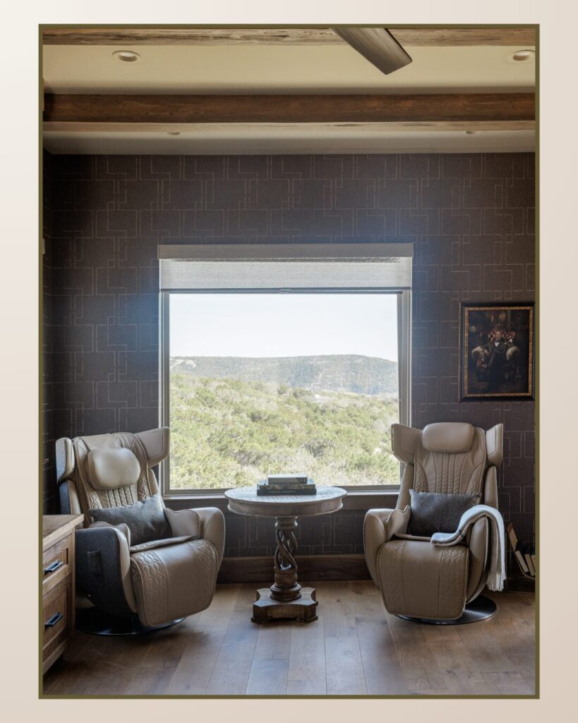 Two stylish beige chairs face a circular table by a large window, offering a view of green hills and a rustic painting on the wall.