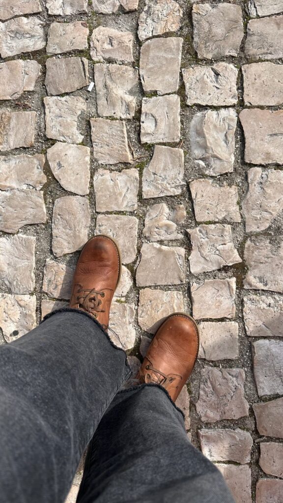 Brown leather shoes on a cobblestone path, featuring light gray stones with patches of green moss.