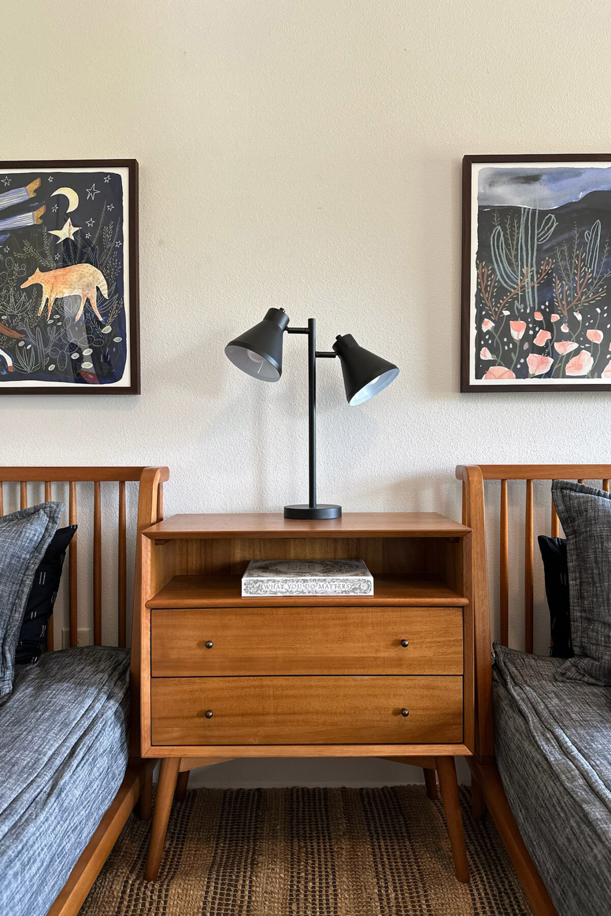 Mid-century wooden nightstand flanked by two gray-textured beds, with black lamps and colorful framed artwork above.