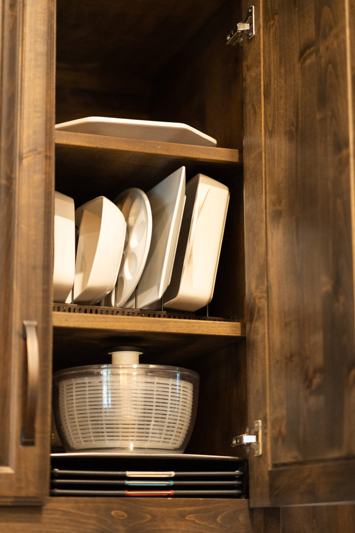 Neatly organized wooden cabinet showcasing white plates, a clear salad spinner, and colorful stacked dishes, emphasizing functional design.