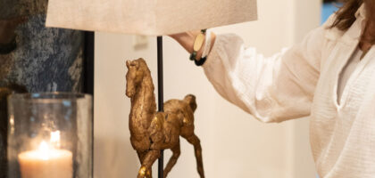 A woman adjusts a stylish lamp featuring a bronze horse, accompanied by a lit pillar candle and stacked books on a wooden table.