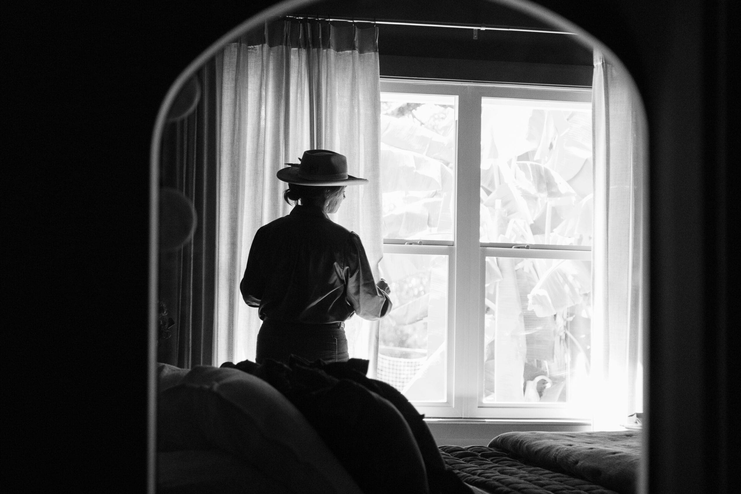 Tiffany is wearing a wide brimmed hat while adjusting sheer curtains at a window. The image is in black and white